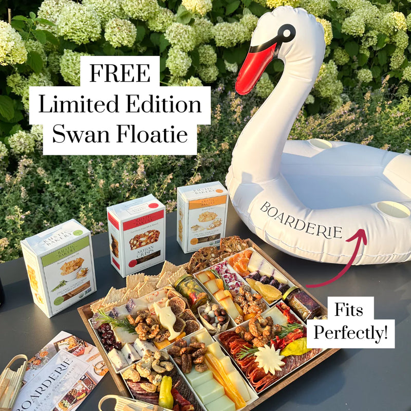 Summer charcuterie board display alongside boxes of artisan crackers & a limited edition swan floatie labeled "Boarderie" on a table outdoors