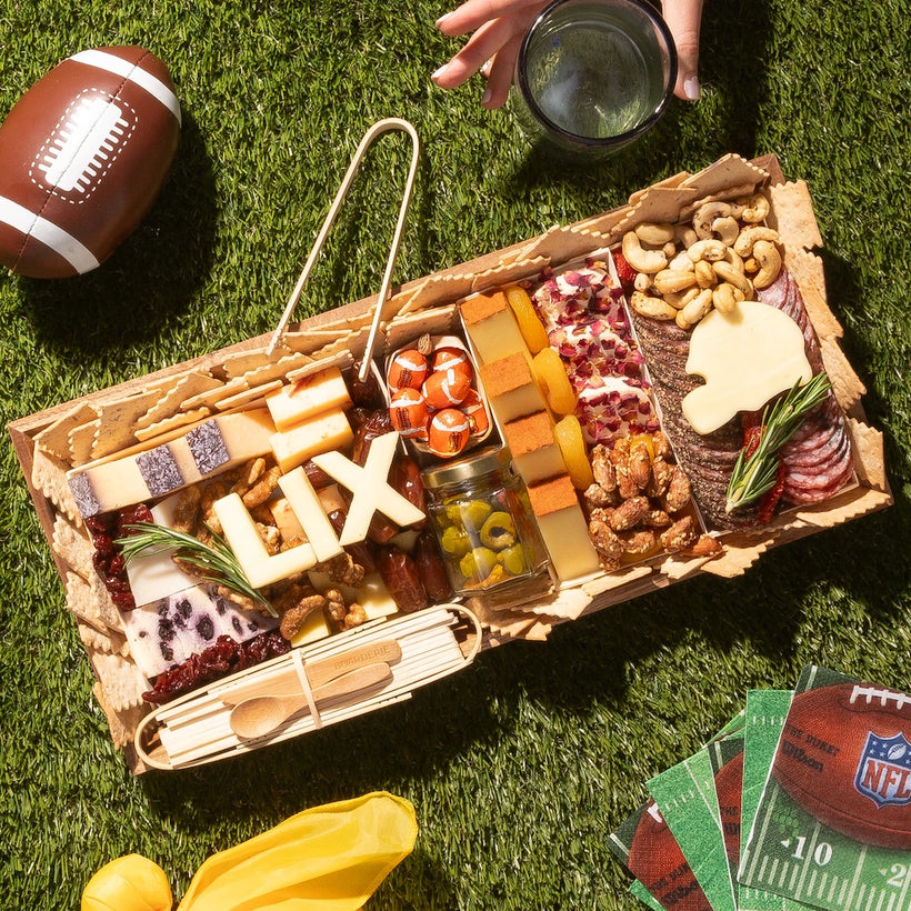 Super Bowl charcuterie board featuring cheeses, meats, crackers, nuts, and football-themed cheese cutouts on a picnic setting.