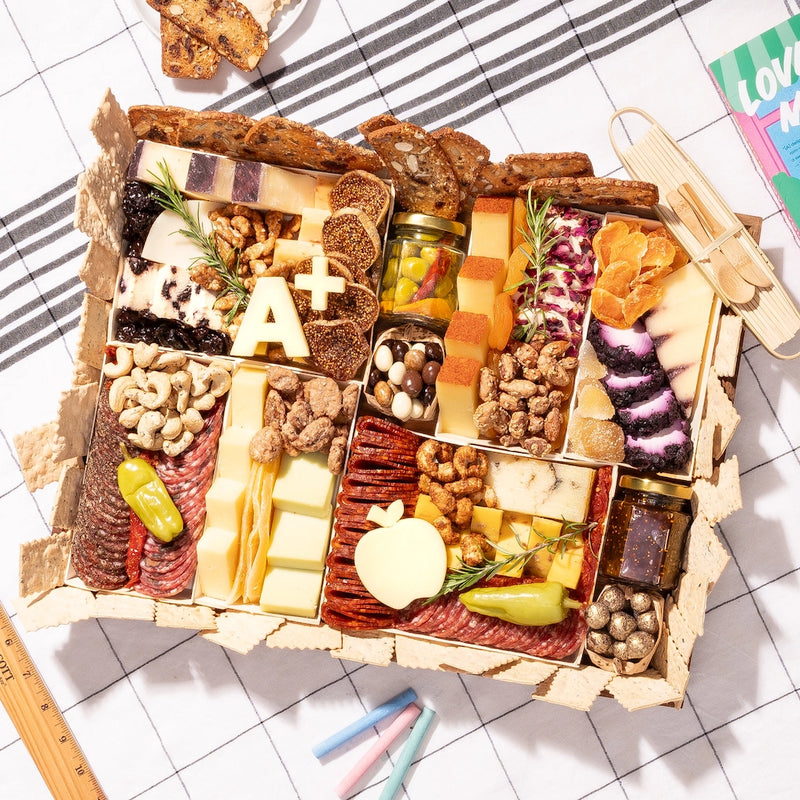 teacher-appreciation charcuterie board with cheese, meats, nuts, an "A+" cutout, & apple-shaped cheese on a checkered cloth