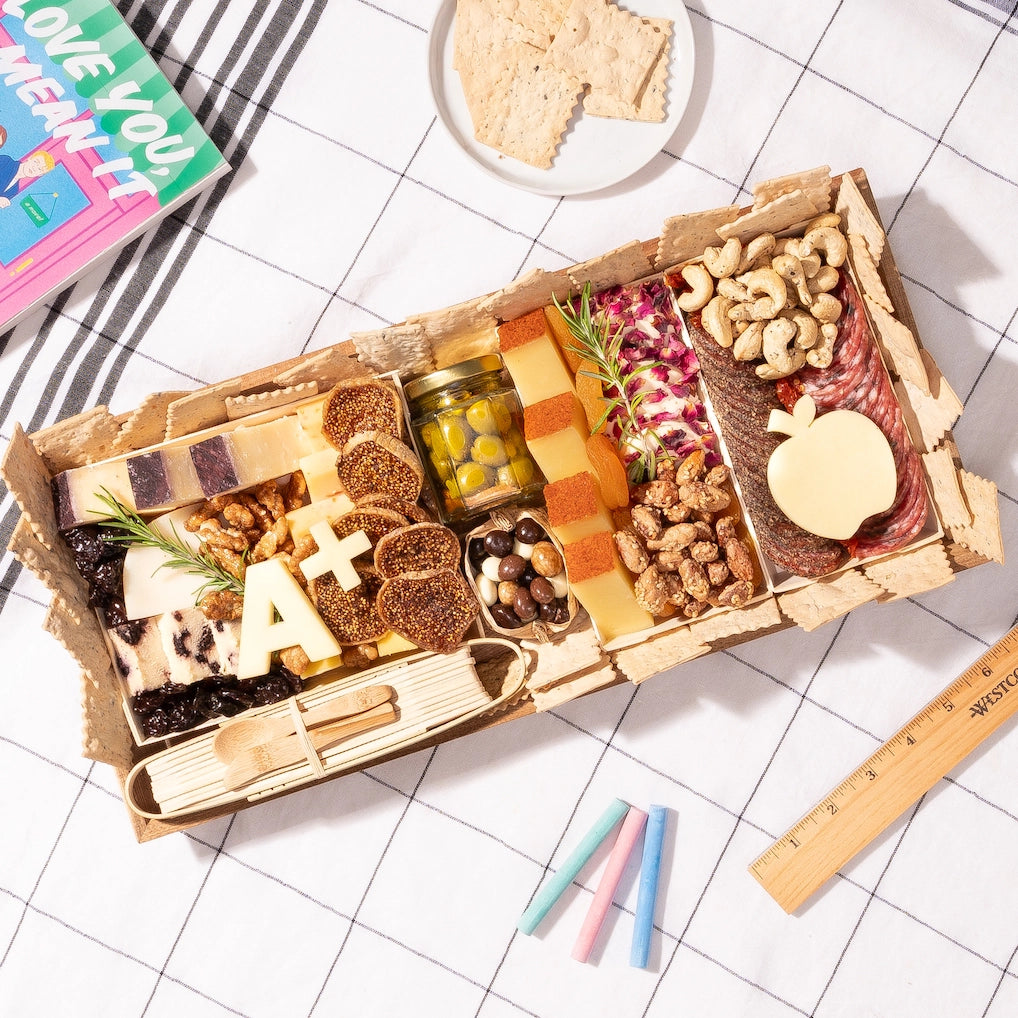 medium teacher-appreciation charcuterie board with cheeses, meats, nuts, apple-shaped cheese, & an "A+" cutout, on a checkered tablecloth with school supplies