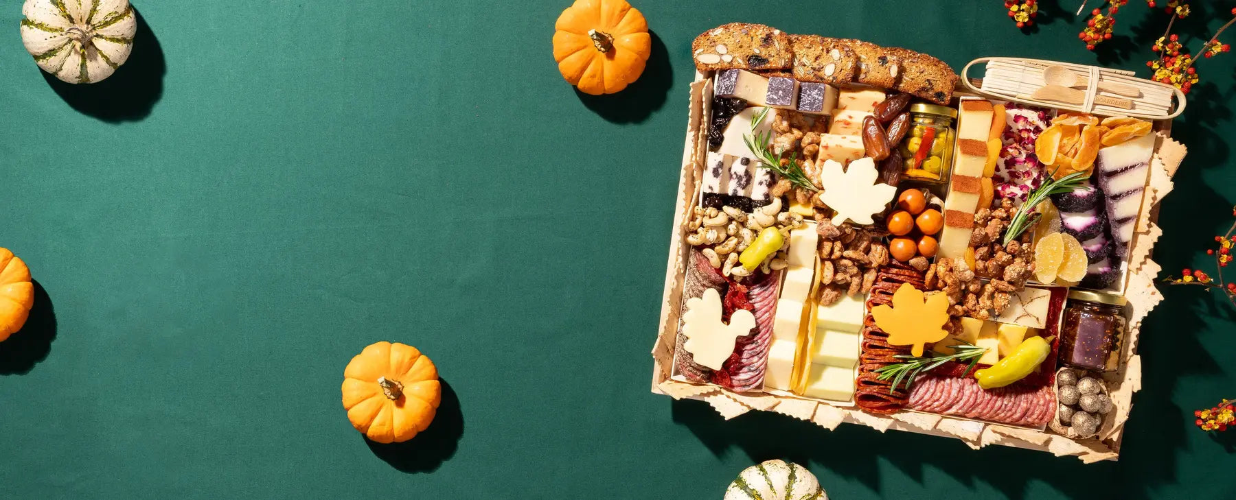 Thanksgiving charcuterie board with assorted cheeses, meats, crackers, dried fruits, and fall-themed decorations on a green tablecloth.