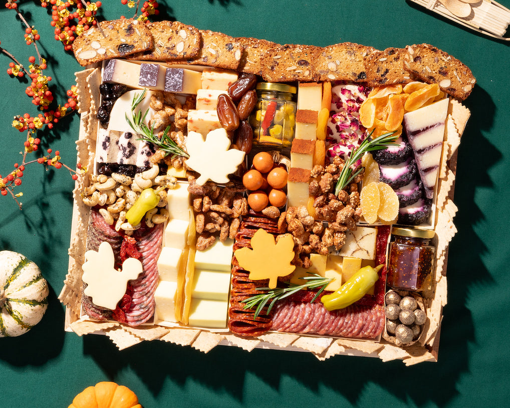 Thanksgiving charcuterie board with assorted cheeses, meats, crackers, dried fruits, and fall-themed decorations on a green tablecloth.