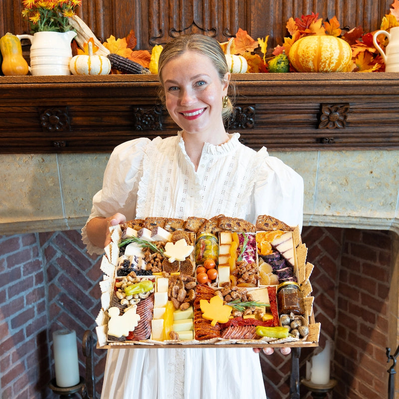 Woman holding a Thanksgiving charcuterie board filled with cheeses, meats, nuts, and fall-themed decorations in front of a festive mantel.