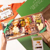 Hands holding a Thanksgiving-themed charcuterie board with cheeses, nuts, dried fruits, & leaf-shaped cheese slices, wrapped in a green Boarderie ribbon.