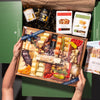 Hands holding an elaborate charcuterie board featuring cheeses, nuts, truffle hot sauce, & assorted snacks, inside a green box