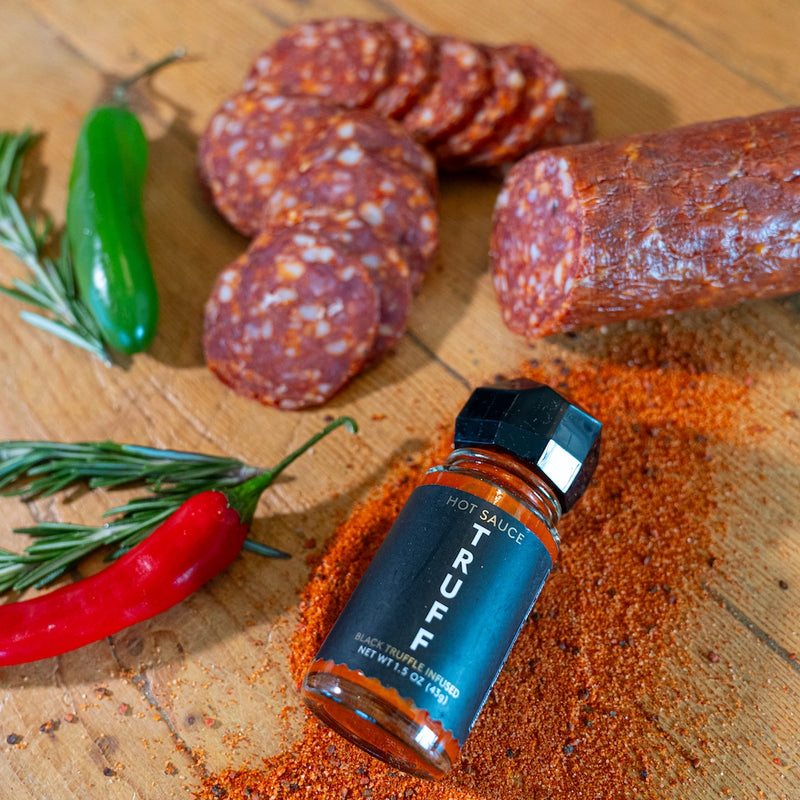 Truffle hot sauce bottle surrounded by sliced salami, chili peppers, & fresh rosemary on a wooden surface