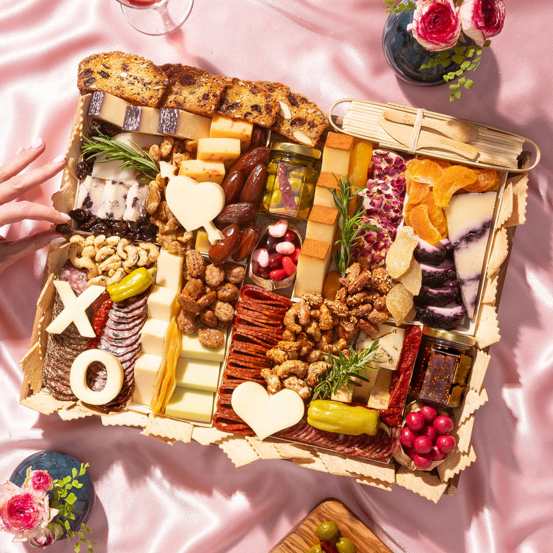 Valentine’s Day charcuterie with gourmet cheeses, meats, nuts, heart-shaped cheese cutouts, and pink accents on a checkered tablecloth.
