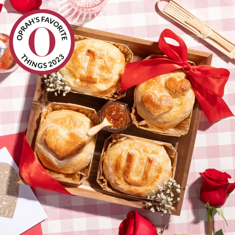 Wooden tray with baked brie pastries spelling "I love you," surrounded by red roses and a jar of jam.