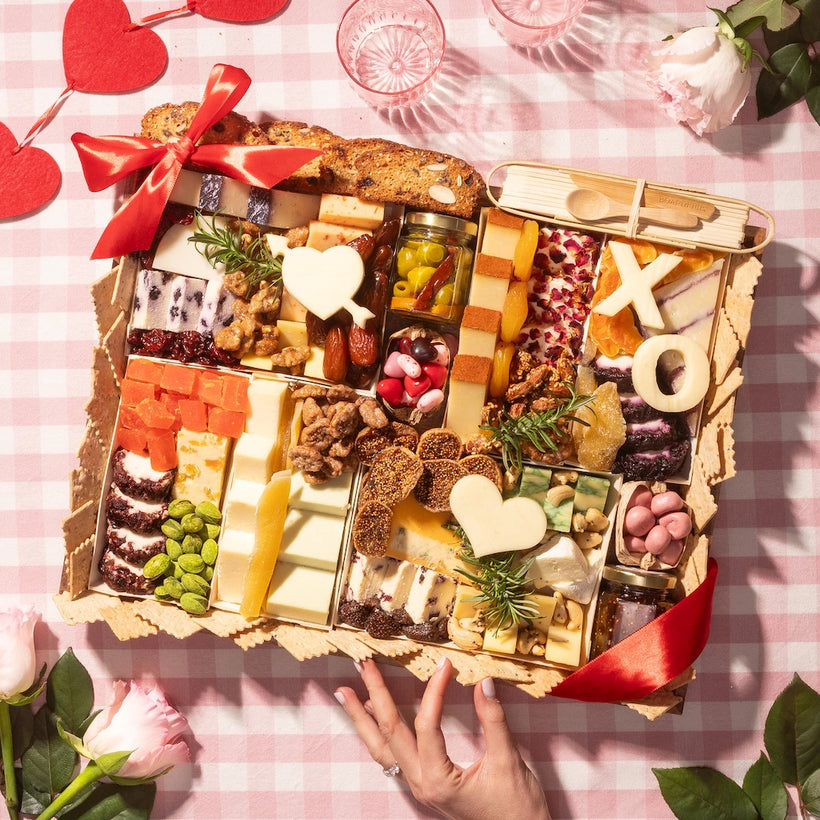 Valentine's Day cheese board with heart-shaped cheeses, nuts, fruits, and crackers on a pink checkered tablecloth with roses.