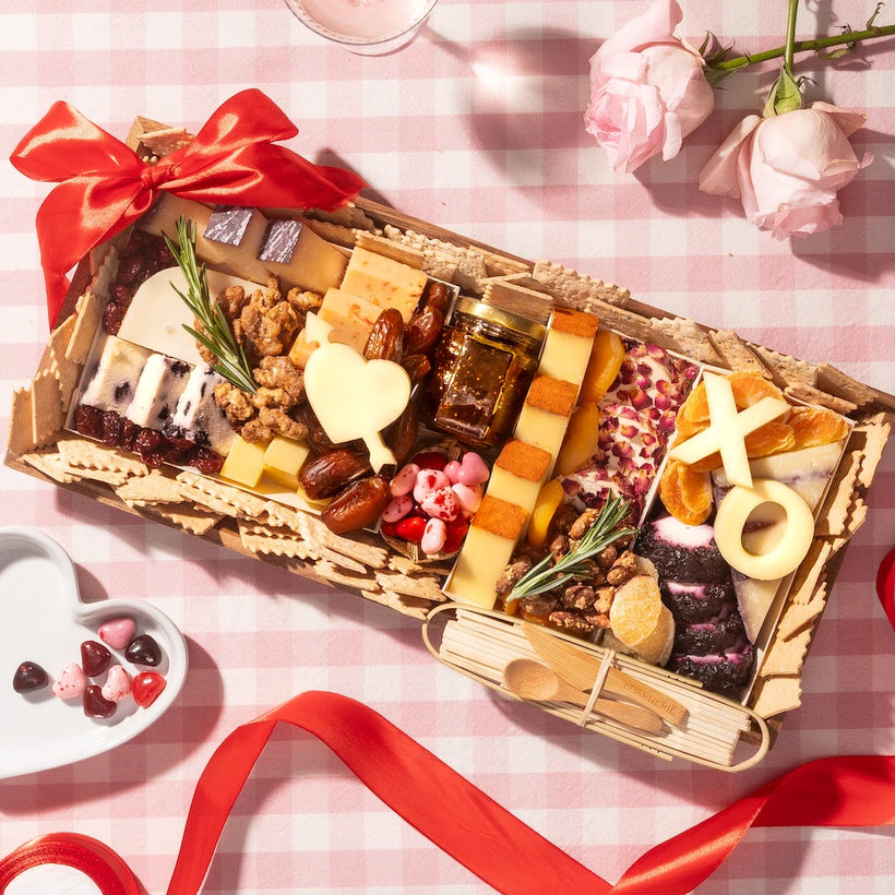 Valentine's Day cheese platter with heart-shaped cheeses, nuts, fruits, and crackers on a pink checkered tablecloth with roses.